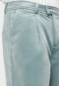 Calças de veludo azul claro com um design plissado, passadores para cinto e bolsos laterais, apresentando uma textura suave e macia.