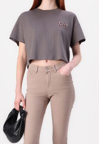 Camiseta gris corta con un logo en el pecho, combinada con jeans beige de cintura alta. El modelo sostiene un bolso negro anudado.