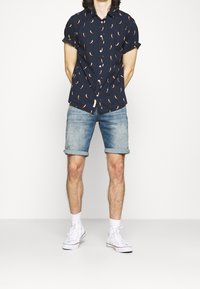 Camisa de manga corta azul marino con estampado de pimientos rojos, combinada con pantalones cortos de mezclilla con dobladillo enrollado, zapatillas blancas y calcetines blancos.