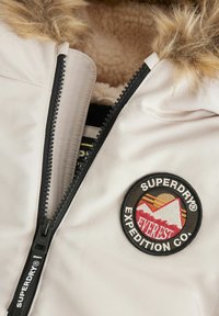 Veste beige clair avec capuche en fourrure, fermeture éclair noire et un patch rond brodé représentant des montagnes et le nom de la marque "Superdry."