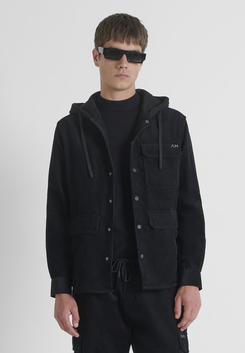 Chaqueta de pana negra con capucha, botones a presión y dos bolsillos frontales. Lleva una camiseta de cuello alto negra debajo y está combinada con pantalones a juego.