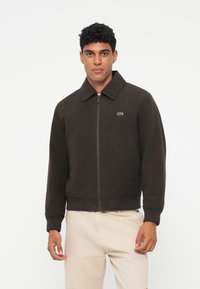 Rudas užtrauktuku užsegamas džemperis su plokščiu apykakle, banguotomis rankovėmis ir apačia, ant krūtinės yra mažas žalias Lacoste logotipas.