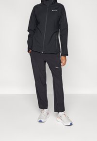 Czarna wodoodporna kurtka zapinana na zamek z przodu, z kieszeniami na zamek i kapturem; w poł�ączeniu z czarnymi spodniami z zamkami oraz szarymi sneakersami.