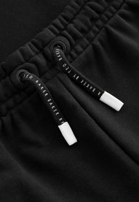 Schwarze Shorts mit Kordelzug aus strukturiertem Stoff, einem gerafften Bund und kontrastierenden weißen Enden an den Kordeln. Logo-Detail sichtbar.