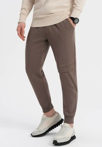 Brune joggerbukser lavet af blødt stof med elastiske manchetter, sidelommer og et strømlinet design, parret med lyse sneakers.