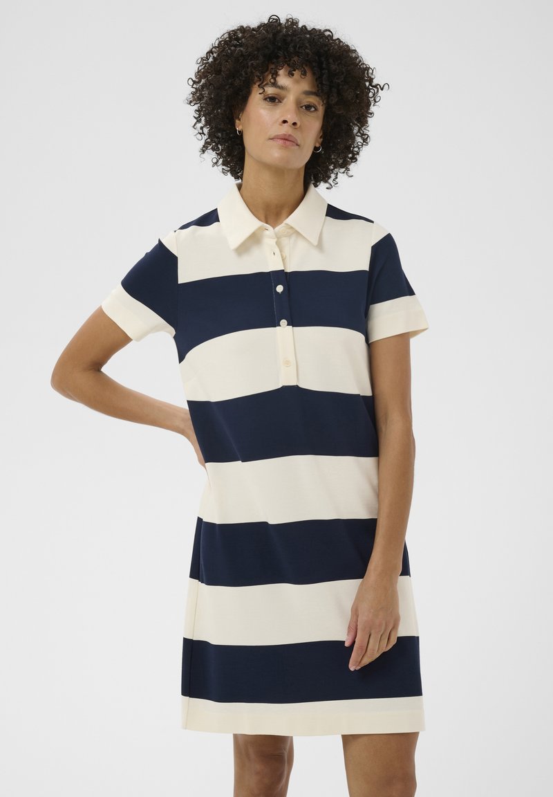 Femme aux cheveux bouclés portant une robe courte à manches courtes, longueur genou, à rayures horizontales bleu marine et blanches, avec un col et des boutons.