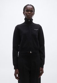 Fekete, cipzáras dzseki texturált anyagból, magas gallérral, két első zsebbel és fehér "tommyjeans" logóval.