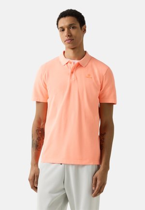 Junger Mann trägt ein pfirsichfarbenes Polohemd mit kurzen Ärmeln und kleinem Logo sowie helle Hosen, stehend vor einem einfarbigen Hintergrund.