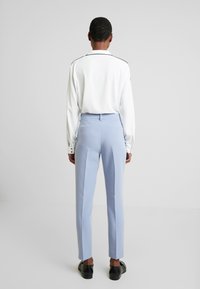 Persona in piedi, di spalle, che indossa una camicia bianca a maniche lunghe con bordi neri, pantaloni su misura azzurri chiari e scarpe eleganti nere.