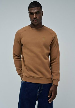 Sweatshirt marron avec un col et des poignets côtelés, arborant un petit logo en patch sur la poitrine. Porté avec un jean en denim bleu foncé.