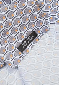 Overhemd met kraag, gemaakt van een witte stof met een herhalend patroon van tennisrackets en oranje tennisballen; zwart label met de tekst: "Elke stap een nieuwe reis."