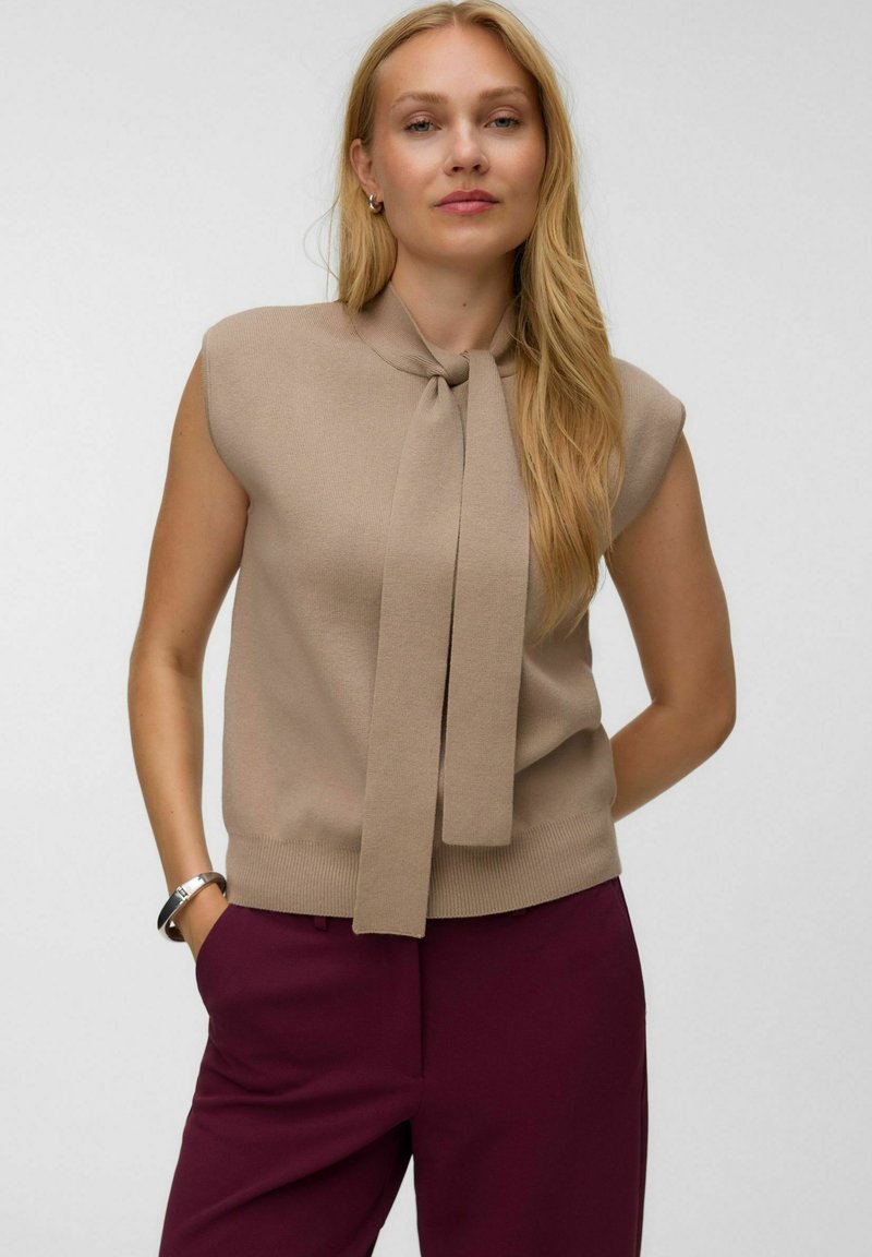 Femme aux longs cheveux blonds portant un top beige sans manches avec un détail noué et un pantalon violet foncé, debout les mains dans les poches.