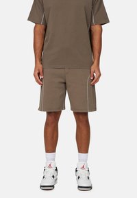 Bruine katoenen t-shirt en bijpassende shorts met witte stiksels, gecombineerd met witte en zwarte sneakers en witte crew-sokken.