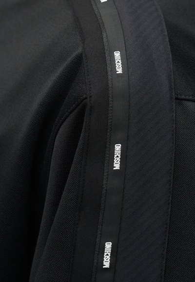 Κοντινό πλάνο μαύρου υφάσματος με υφασμάτινο πάνελ και μαύρη λωρίδα με επαναλαμβανόμενο λευκό κείμενο "MOSCHINO".