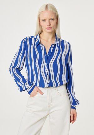 Blonde woman wearing blue and white wavy striped long-sleeve blouse tucked into high-waisted white pants with one hand in pocket.