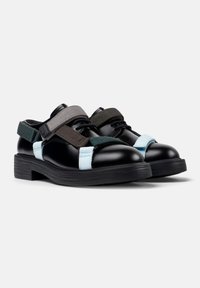 Zapatos de cuero negros con amplias correas elásticas en gris, verde oscuro y azul claro. Suelas gruesas de goma negra, diseño de punta redondeada.