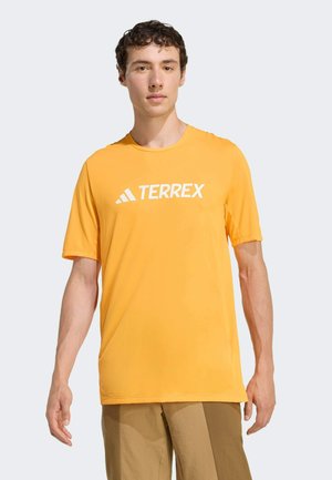 Junger Mann, der ein gelbes Adidas Terrex Kurzarmshirt und zweifarbige braune Hosen trägt, steht vor einem schlichten hellen Hintergrund.
