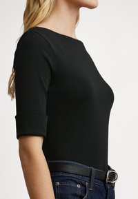 Μαύρο εφαρμοστό τοπ με μανίκια τριών τετάρτων, στρογγυλή λαιμόκοψη, λεία υφή, σε συνδυασμό με σκουρόχρωμο τζιν και μαύρη ζώνη.