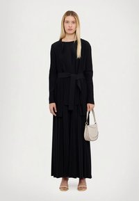 Μαύρο μακρύ φόρεμα με πτυχώσεις και ζώνη, μακριά μανίκια, στρογγυλή λαιμόκοψη και πλαϊνή σχισμή. Αξεσουάρ περιλαμβάνουν μία τσάντα σε ανοιχτό μπεζ και σανδάλια.