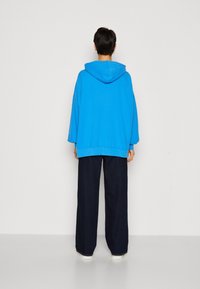 Felpa con cappuccio blu a vestibilità ampia e maniche lunghe, abbinata a pantaloni larghi scuri. Tessuto liscio con polsini e orlo a costine.