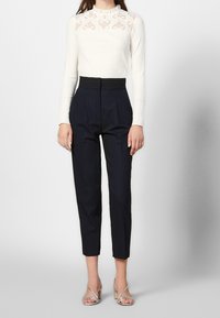 En person står klädd i en snug vit topp med spetsdetaljer och högmidjade marinblå byxor, kombinerat med silverfärgade klacksandaler.