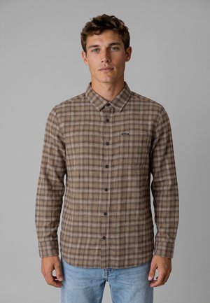 Giovane uomo con capelli corti e ricci che indossa una camicia marrone a quadri con bottoni e jeans azzurri, in piedi contro uno sfondo grigio uniforme.