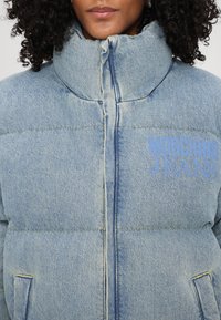 Gaiši zila džinsa pufīga jaka ar augstu apkakli, šūtu šuvju un zilu "Moschino Jeans" logo. Ievērojamas lielas kabatas un teksturēta auduma. 