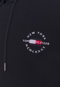Sudadera negra de mezcla de algodón con un logo bordado circular que incluye "NUEVA YORK", "TOMMY HILFIGER" y "MCMLXXXV", con acentos en rojo y azul.