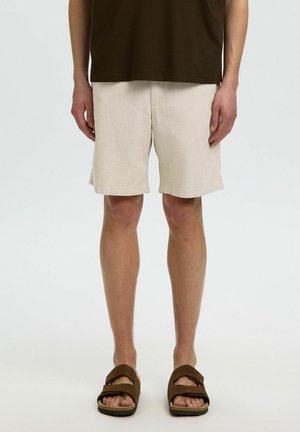 Homme portant un short beige, un haut marron foncé et des sandales ouvertes marron, debout avec les mains détendues le long du corps sur un fond blanc uni.