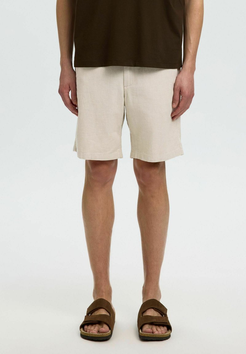 Homme portant un short beige, un haut marron foncé et des sandales ouvertes marron, debout avec les mains détendues le long du corps sur un fond blanc uni.