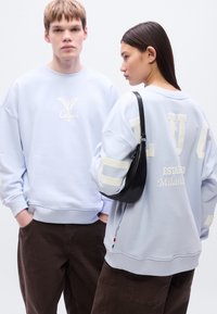 Ανοιχτό μπλε φαρδιά φούτερ με λογότυπα μπροστά και πίσω, συνδυασμένα με καφέ παντελόνια. Η υφή του φούτερ είναι μαλακή και περιλαμβάνει αντίθετες λεπτομέρειες.