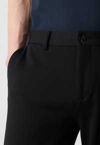 Czarne spodnie na miarę wykonane z gładkiego materiału, z bocznymi kieszeniami, płaskim przodem i zapięciem na guzik w pasie z pętlą.