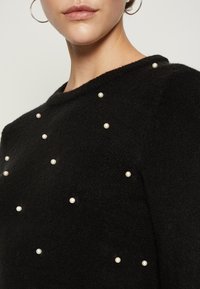 Svart stickad tröja med rund halsringning, prydd med spridda vita pärldekorationer över framsidan. Mjuk textur, avslappnad passform.