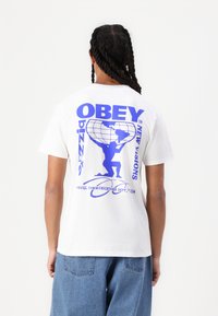 Hvit bomulls T-skjorte med et blått grafisk motiv på baksiden, som viser en figur som holder en klode med teksten "OBEY" og "NEW VISIONS".
