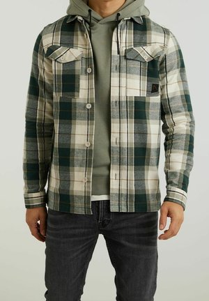 Uomo che indossa una camicia a quadri verde e bianca con bottoni sopra una felpa verde con cappuccio e jeans scuri, in piedi contro uno sfondo semplice.