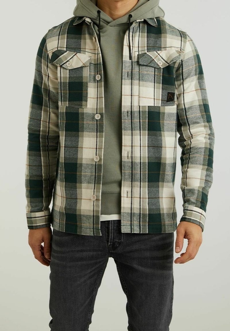 Homme portant une chemise à carreaux verte et blanche boutonnée par-dessus un sweat à capuche vert avec un jean foncé, debout devant un fond uni.
