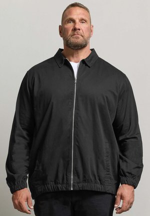 Homme aux cheveux courts et à la barbe, portant une veste noire zippée avec des poignets et un col élastiques, debout devant un fond clair uni.