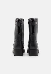 Un par de botines de cuero negro con tacones de bloque y correas traseras, mostrados desde la parte trasera sobre un fondo blanco.