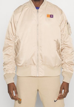 Veste bomber beige avec des poignets et un ourlet côtelés, fermeture éclair à l'avant, poches latérales et un écusson avec des rayures et un blason sur la poitrine.