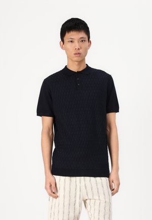 Polo en maille bleu marine avec un motif en diamants texturé, doté d'un col noir et d'une patte de boutonnage à trois boutons. Porté avec un pantalon clair à rayures.