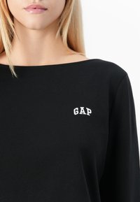 Svart långärmad topp av bomull med vid halsringning. Har en liten vit broderad "GAP"-logotyp på den övre vänstra sidan.