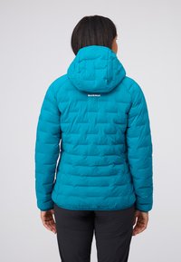 Teal vadderad jacka med huva och quiltat mönster. Har en logotyp på nacken och elastiska ärmslut. Strukturerad tyg och figursydd form.