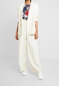 Blazer bianco doppiopetto sopra una maglietta a stampa floreale con rose rosse su sfondo scuro, abbinato a pantaloni bianchi larghi e sneakers bianche.