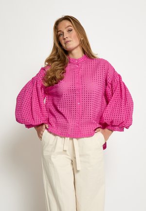 Helles pinkes, langärmeliges Blusenoberteil mit Puffärmeln, das ein strukturiertes Design mit ausgeschnittenen Punkten und eine Knopfleiste vorne aufweist. Kombiniert mit hellen Hosen.