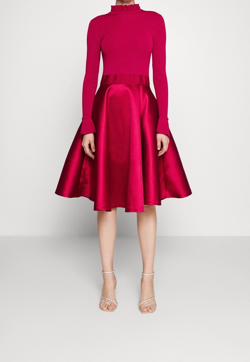 Femme portant un haut ajusté à manches longues fuchsia avec un col à volants et une jupe midi évasée en satin, associée à des sandales à talons aiguilles à brides.