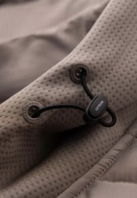 Ljusbrun textil med en texturerad, småprickig mönstring. Innehåller svarta spännen och snoddar för justering, med logotypdetaljer på hårdvaran.