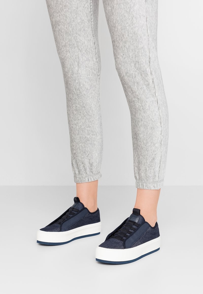 Ben som bär ljusgråa sweatpants och marinblå plattformssneakers med vita sulor på en neutral ljus bakgrund.