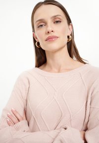 Hellrosa gestrickter Pullover mit strukturiertem Rautenmuster, geripptem Ausschnitt und langen Ärmeln. Goldene Kreolenohrringe setzen einen akzentuierenden Akzent.