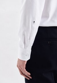 Camicia bianca in cotone con polsini abbottonati, caratterizzata da un piccolo ricamo di una rosa nera sul polsino e una finitura testurizzata. Pantaloni neri visibili.