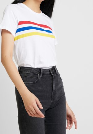 Kobieta w białym t-shircie z czerwonymi, niebieskimi i żółtymi zakrzywionymi paskami, w połączeniu z wysokim stanem wyblakłych czarnych jeansów.